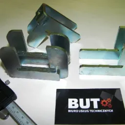 Metalowe elementy spawane lub obrabiane, pomiar suwmiarką elektroniczną, wizytówka Biura Usług Technicznych w tle.