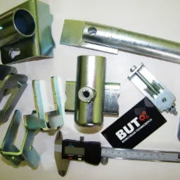 Metalowe elementy po obróbce, suwmiarka cyfrowa i wizytówka Biura Usług Technicznych na białym tle.