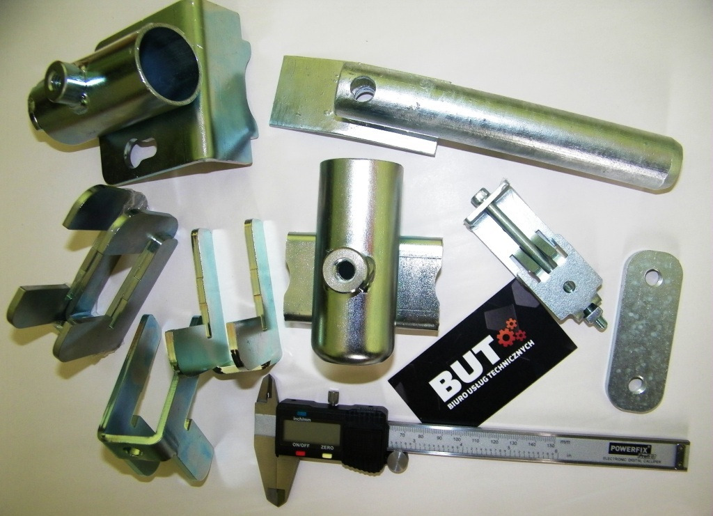 Metalowe elementy po obróbce, suwmiarka cyfrowa i wizytówka Biura Usług Technicznych na białym tle.