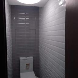 J&K Group Oleksii Kharchenko - Wąska toaleta wykończona szarymi i białymi, błyszczącymi płytkami typu metro, z widoczną zabudową nad stelażem oraz oświetleniem sufitowym typu plafon.