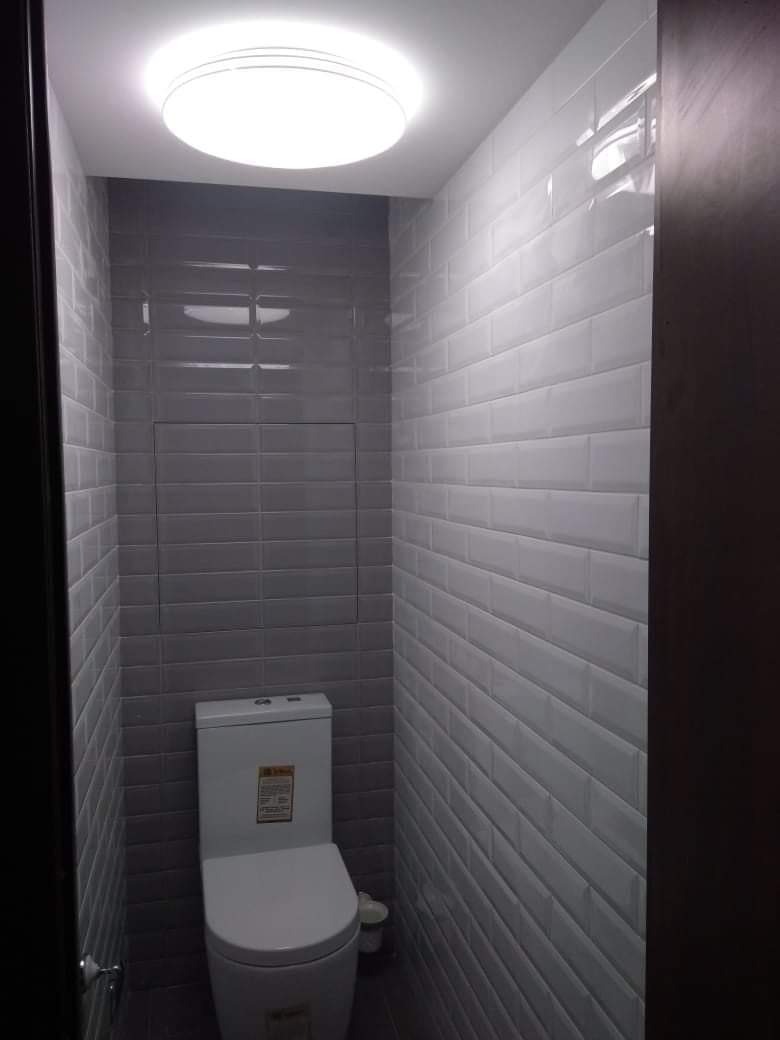 Wąska toaleta wykończona szarymi i białymi, błyszczącymi płytkami typu metro, z widoczną zabudową nad stelażem oraz oświetleniem sufitowym typu plafon.