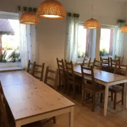 Jasne wnętrze stołówki z drewnianymi stołami i krzesłami, oświetlone wiklinowymi lampami wiszącymi. Za oknami widoczny budynek z czerwonym dachem i zielenią.