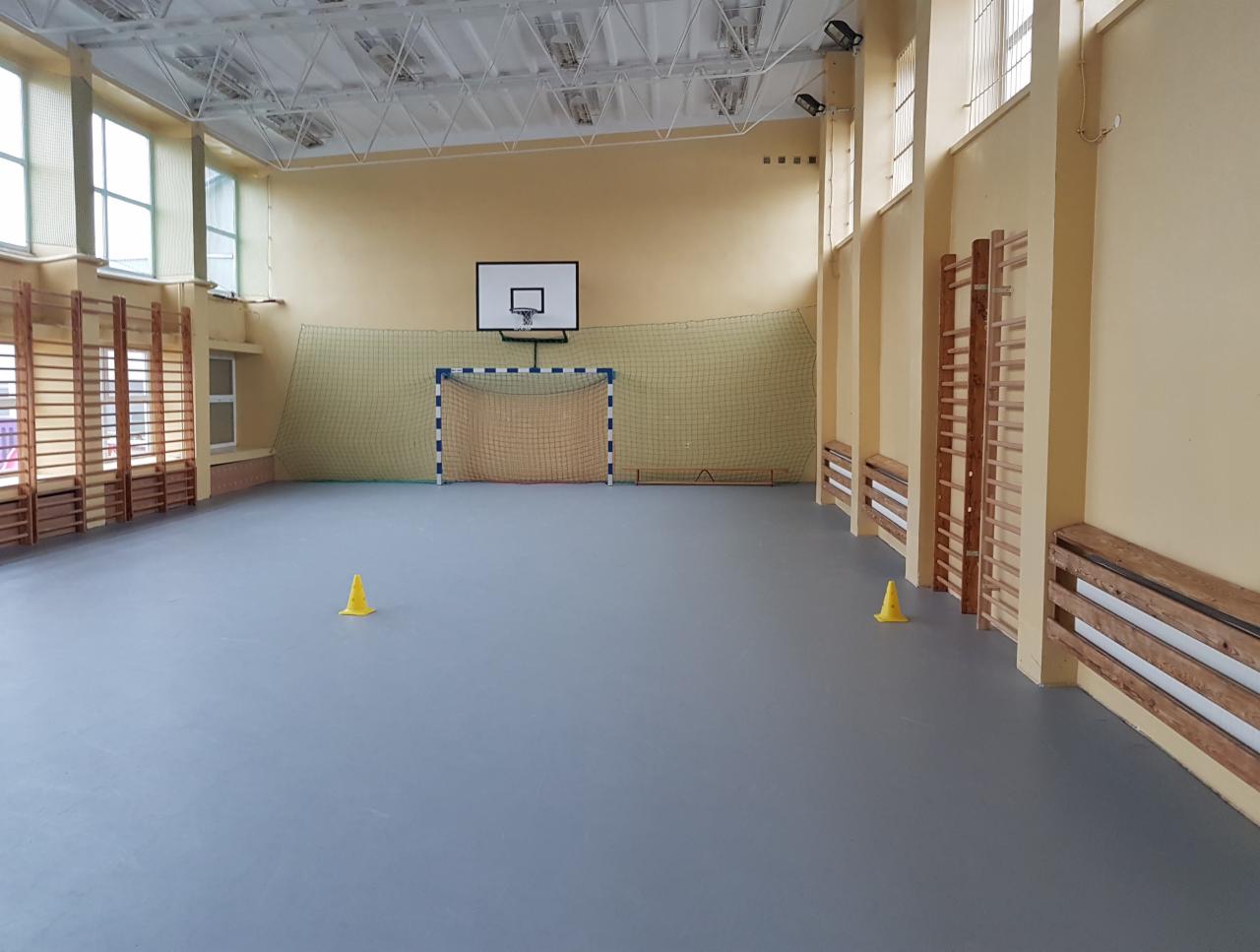 Wnętrze sali gimnastycznej z drabinkami, bramką do piłki ręcznej, koszem do koszykówki i dwoma żółtymi pachołkami na szarej podłodze.