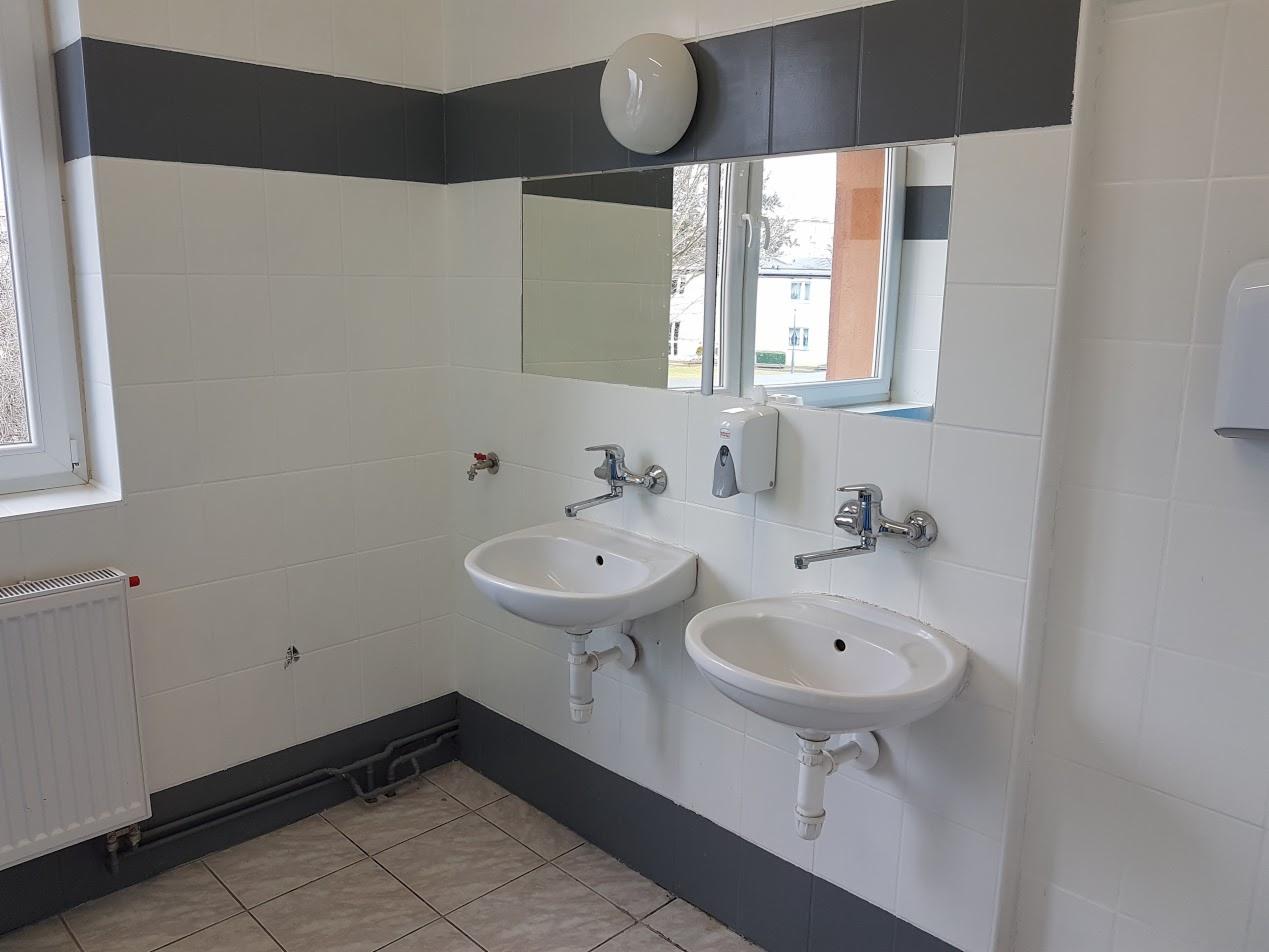 Umywalnia z dwoma białymi umywalkami, chromowanymi kranami, dozownikiem mydła i lustrem. Widoczny grzejnik, białe płytki z szarym pasem na ścianach i okno z widokiem na budynek.