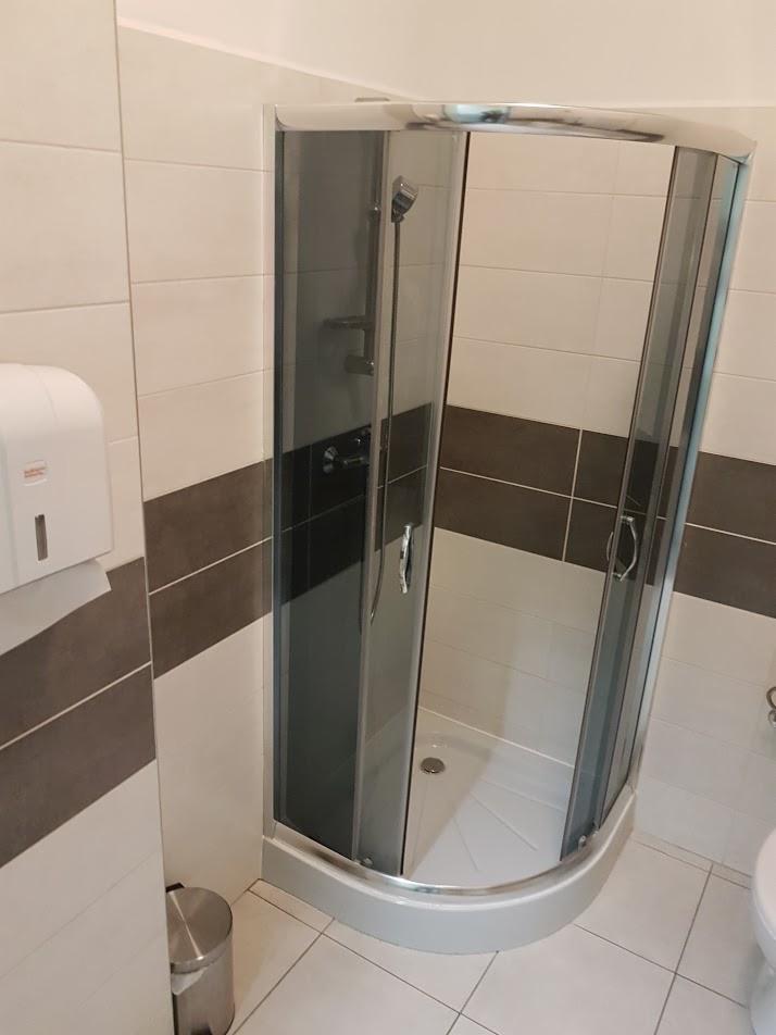 Narożna kabina prysznicowa z chromowanymi profilami i szarymi szybami w łazience z beżowo-brązowymi płytkami, widoczny dozownik mydła i kosz na śmieci.