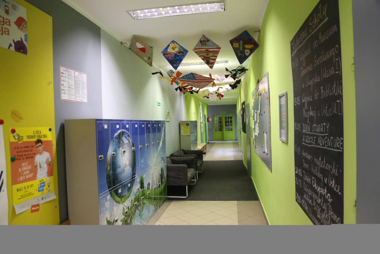 Wnętrze szkolnego korytarza z szafkami z motywem Ziemi, tablicą informacyjną z kalendarzem wydarzeń szkolnych i dekoracjami w postaci latawców.