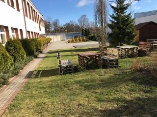 Teren rekreacyjny w Ustce z meblami ogrodowymi wykonanymi z palet, trawnik, budynek w tle, słoneczny dzień.