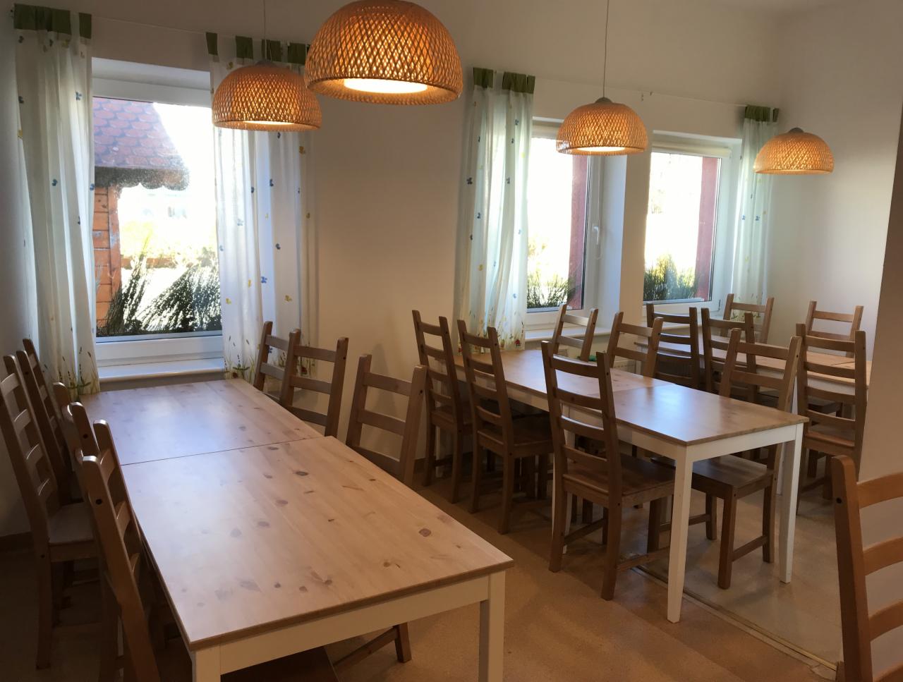 Jasne wnętrze stołówki z drewnianymi stołami i krzesłami, oświetlone wiklinowymi lampami wiszącymi. Za oknami widoczny budynek z czerwonym dachem i zielenią.