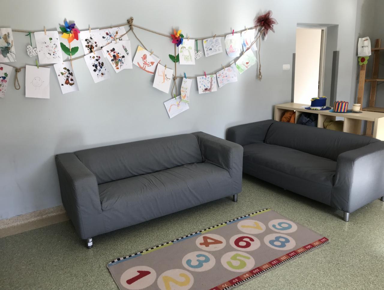 Pokój zabaw z szarymi sofami, dywanem z cyframi i dziecięcymi rysunkami zawieszonymi na sznurku z klamerkami.