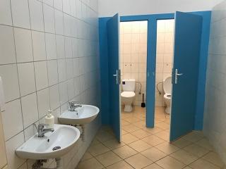 Wnętrze publicznej toalety z dwoma umywalkami po lewej stronie i dwoma kabinami WC z niebieskimi drzwiami po prawej stronie, wyłożone kafelkami w jasnych kolorach.