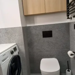 Nowoczesna łazienka z szarą terazzo glazurą, wiszącą toaletą, pralką Samsung i drewnianą szafką nad nią.