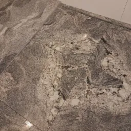 Ułożona pod kątem podłoga z płytek imitujących kamień w odcieniach szarości i beżu, z widoczną fugą i fragmentem białej ściany w tle.