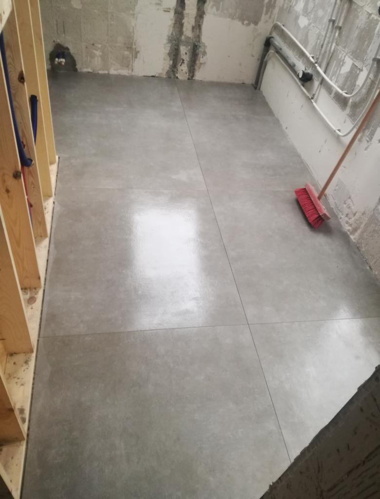 Świeżo wylana, szara posadzka w pomieszczeniu w stanie surowym, widoczne fragmenty ścian bez wykończenia, rury przy suficie i drewniana konstrukcja szkieletowa ściany.