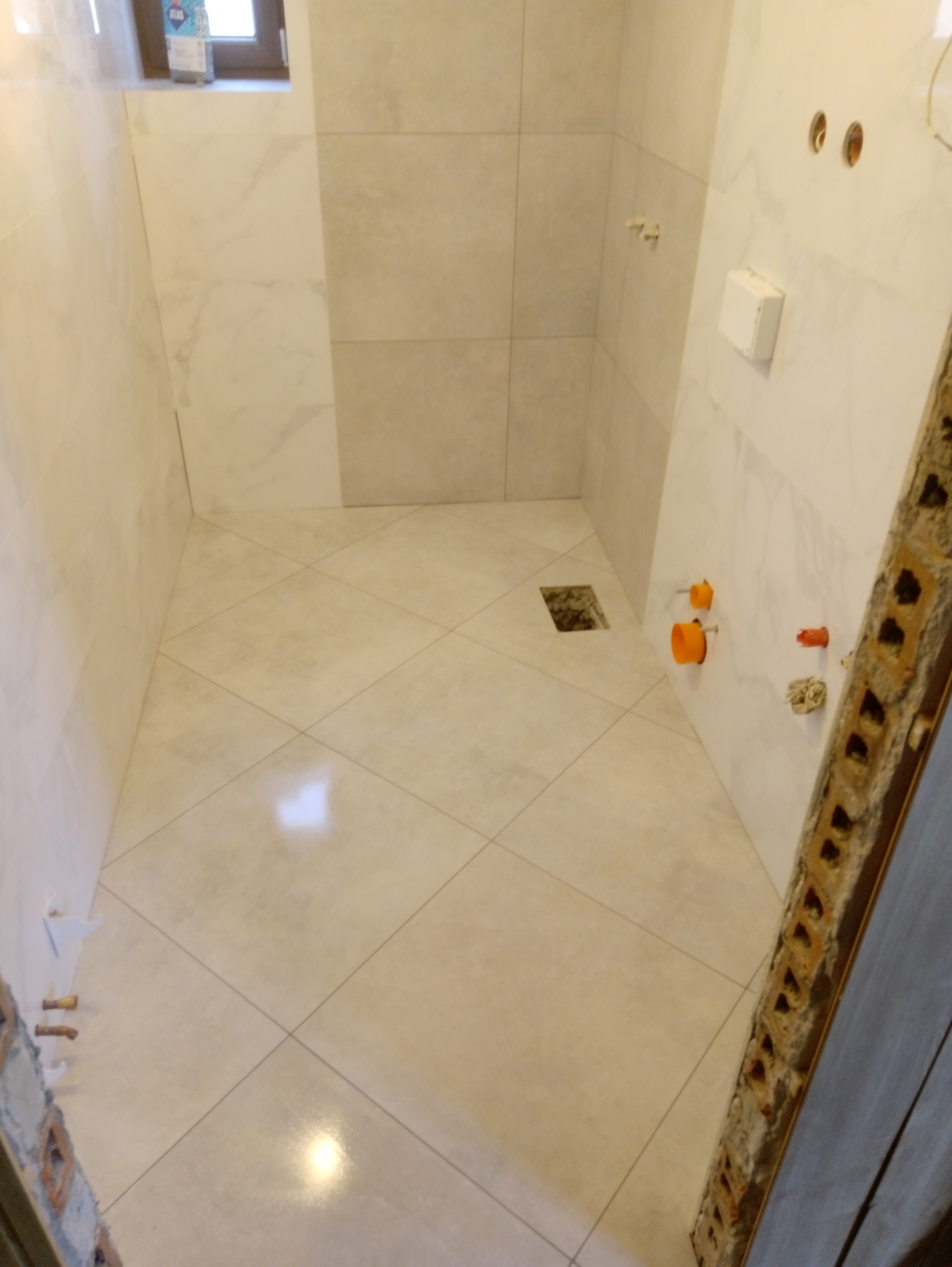 Wnętrze łazienki w trakcie prac wykończeniowych, z ułożonymi jasnymi płytkami podłogowymi w karo, częściowo wykończonymi ścianami z widocznymi instalacjami hydraulicznymi i elektrycznymi...