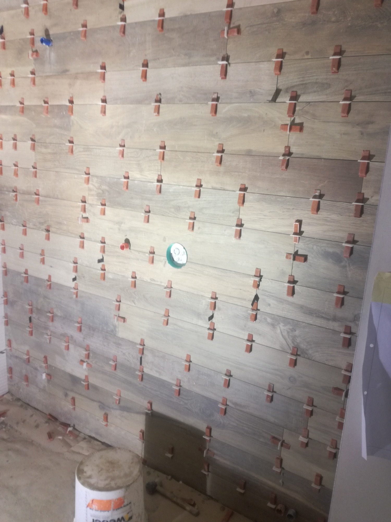 Montaż płytek imitujących drewno na ścianie, z użyciem krzyżyków dystansowych, widoczny fragment instalacji wodnej i wiadro po kleju.