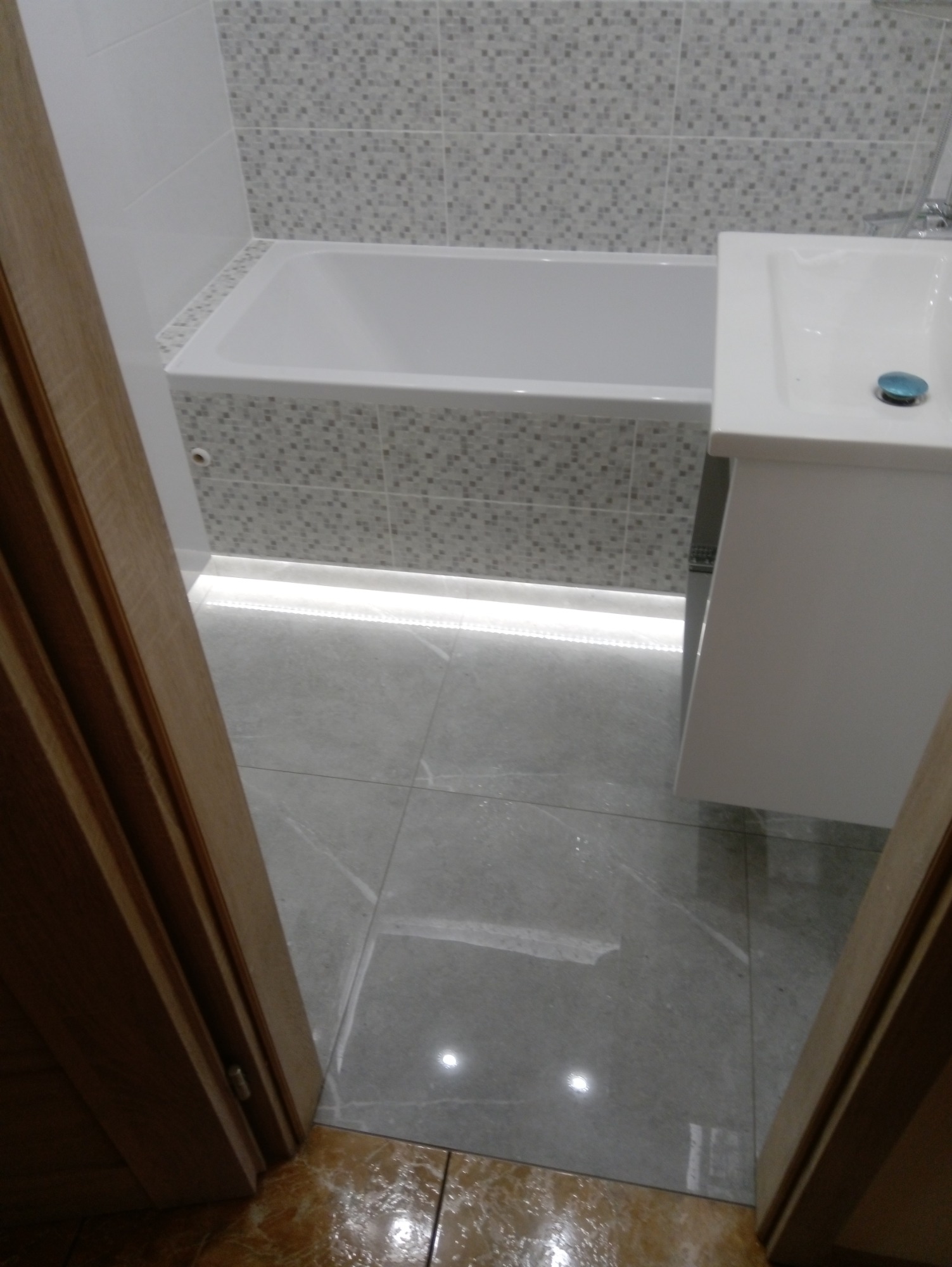 Widok zza uchylonych drzwi na wykończoną łazienkę z wanną obudowaną mozaiką, umywalką z szafką i szarą podłogą z połyskiem, oświetloną listwą LED pod wanną.