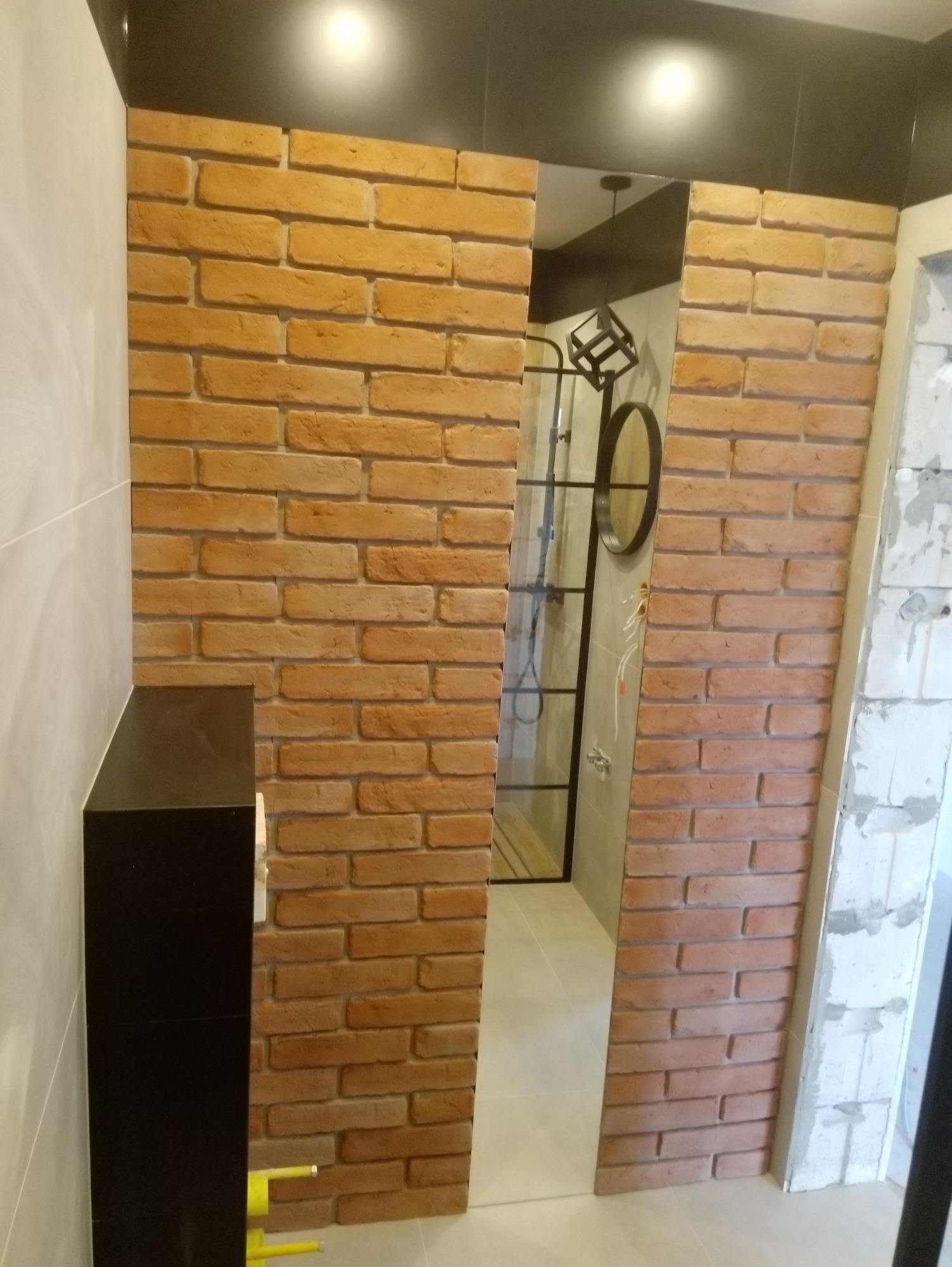 Wykończenie łazienki z dekoracyjną ścianą z cegły, widoczne elementy wyposażenia: kabina prysznicowa z czarną ramą, okrągłe lustro i geometryczna lampa wisząca.
