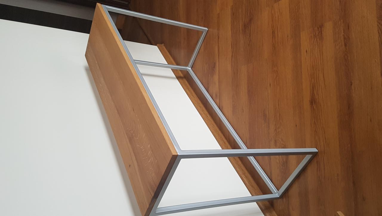 Stelaż drewnianego blatu z metalowym obramowaniem, widoczny na tle białej ściany i drewnopodobnej podłogi.