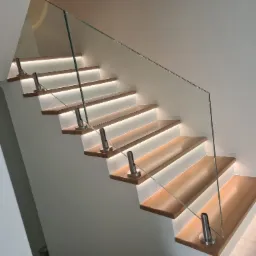 Nowoczesne schody drewniane z podświetleniem LED i szklaną balustradą mocowaną na metalowych słupkach. Minimalistyczny design i jasne wnętrze.