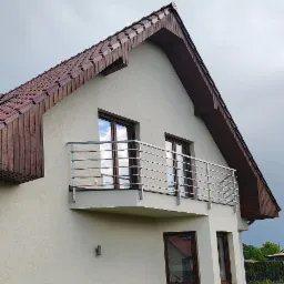 Elewacja domu z balkonem i nowoczesną, metalową balustradą z poziomych prętów. Widok z dołu na fragment budynku z brązową stolarką okienną i drewnianym wykończeniem dachu.