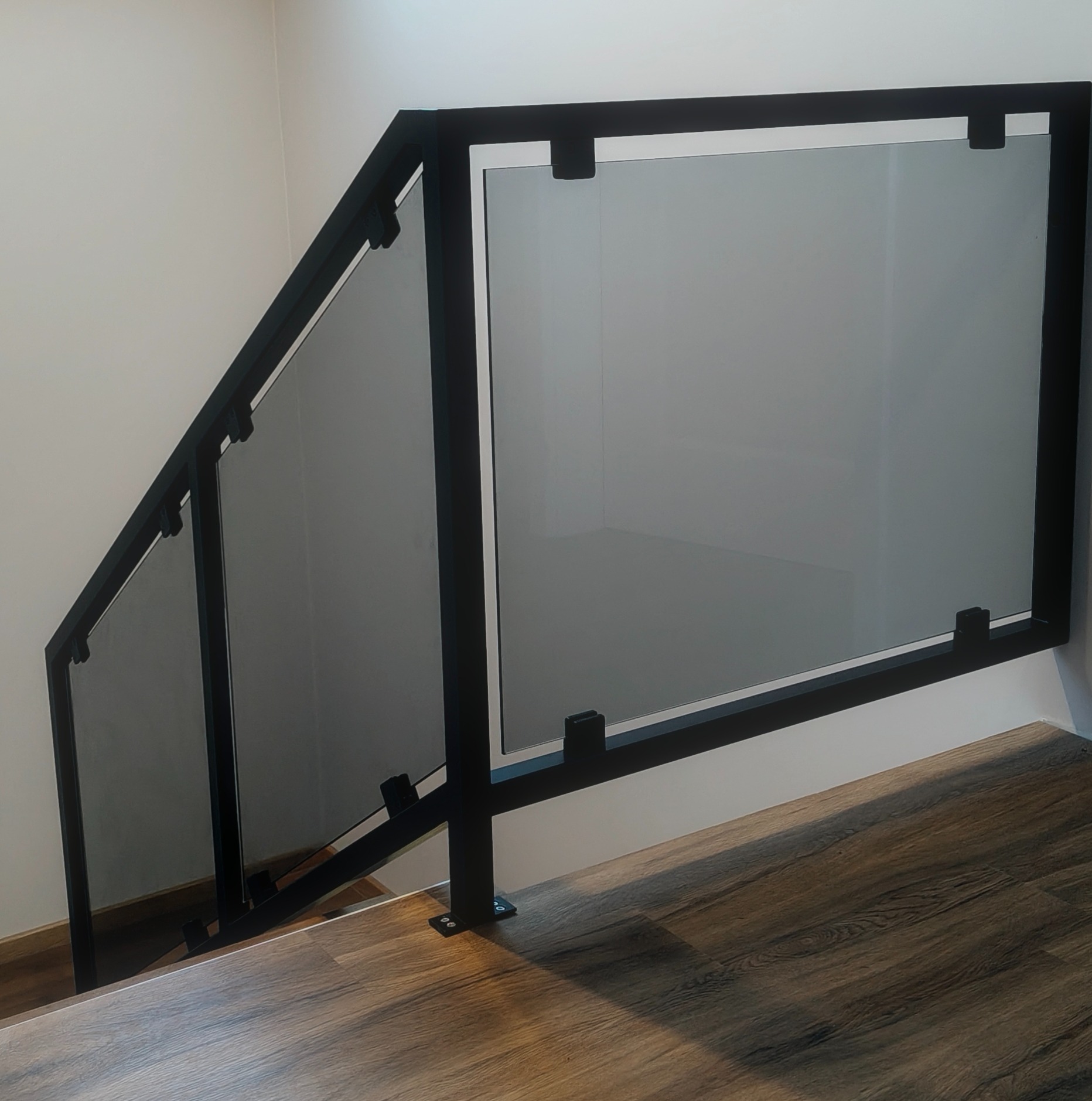 Szklana balustrada schodowa z czarną, metalową ramą i mocowaniami. Widok z dołu, ukazujący drewnopodobne wykończenie schodów i minimalistyczny design.