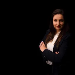 Portret kobiety w białej koszuli i ciemnej marynarce na czarnym tle, z rękami skrzyżowanymi na piersiach, patrzącej w obiektyw z lekkim uśmiechem.