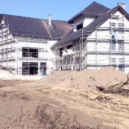 Budynek w trakcie budowy z widoczną konstrukcją dachu, rusztowaniami i stertą ziemi na pierwszym planie.