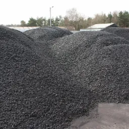 Kupy ciemnego, drobnego węgla na placu składowym, widoczne budynki magazynowe i drzewa w tle.