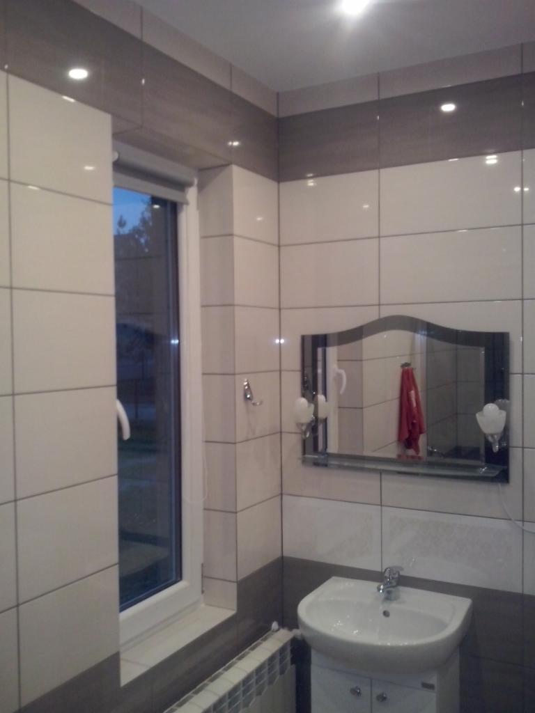 Wykończona łazienka z białymi i szarymi płytkami, umywalką z szafką, lustrem z oświetleniem i grzejnikiem pod oknem.