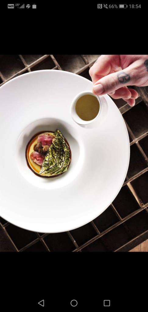 Elegancko podane danie z mięsem, sosem i kapustą na białym talerzu, z dodatkowym sosem w małym dzbanku trzymanym przez wytatuowaną dłoń, na tle kratownicy.