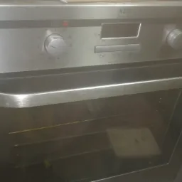 Metalowy piekarnik marki AEG z widocznym panelem sterowania i uchwytem, obok leży wiertarka i kabel zasilający.