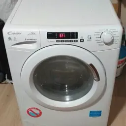 Biała pralka Candy Alise z wyświetlaczem pokazującym błąd E21, umieszczona na drewnianej podłodze, z widocznymi przedmiotami w tle.