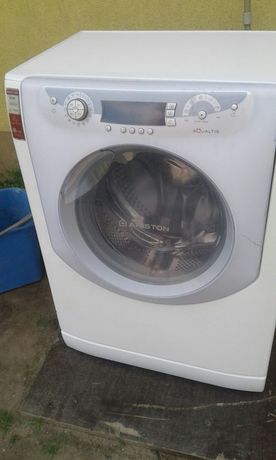 Biała pralka Ariston Aqualtis z widocznym panelem sterowania i bębnem, stojąca na szarym podłożu, z fragmentem niebieskiego wiadra w tle.