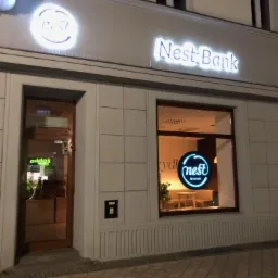Fasada budynku Nest Bank z podświetlanymi neonami logo i nazwą, widziana z zewnątrz o zmierzchu.