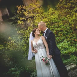 Panna młoda w koronkowej sukni i welonie trzyma bukiet różowych kwiatów, pan młody w bordowej marynarce całuje ją w czoło na tle zieleni.