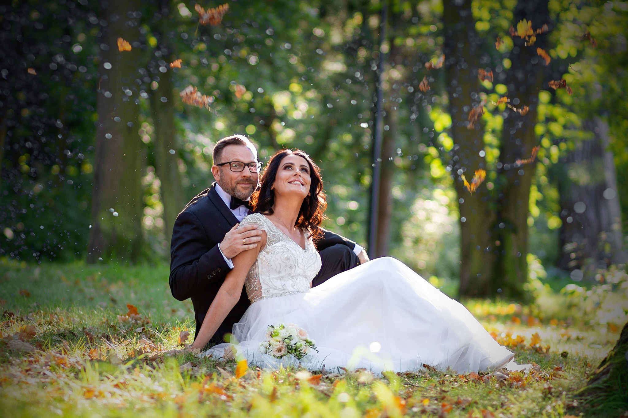 Para młoda siedząca na trawie w lesie, panna młoda w białej sukni z bukietem, pan młody w czarnym garniturze, opadające liście, słoneczne światło.