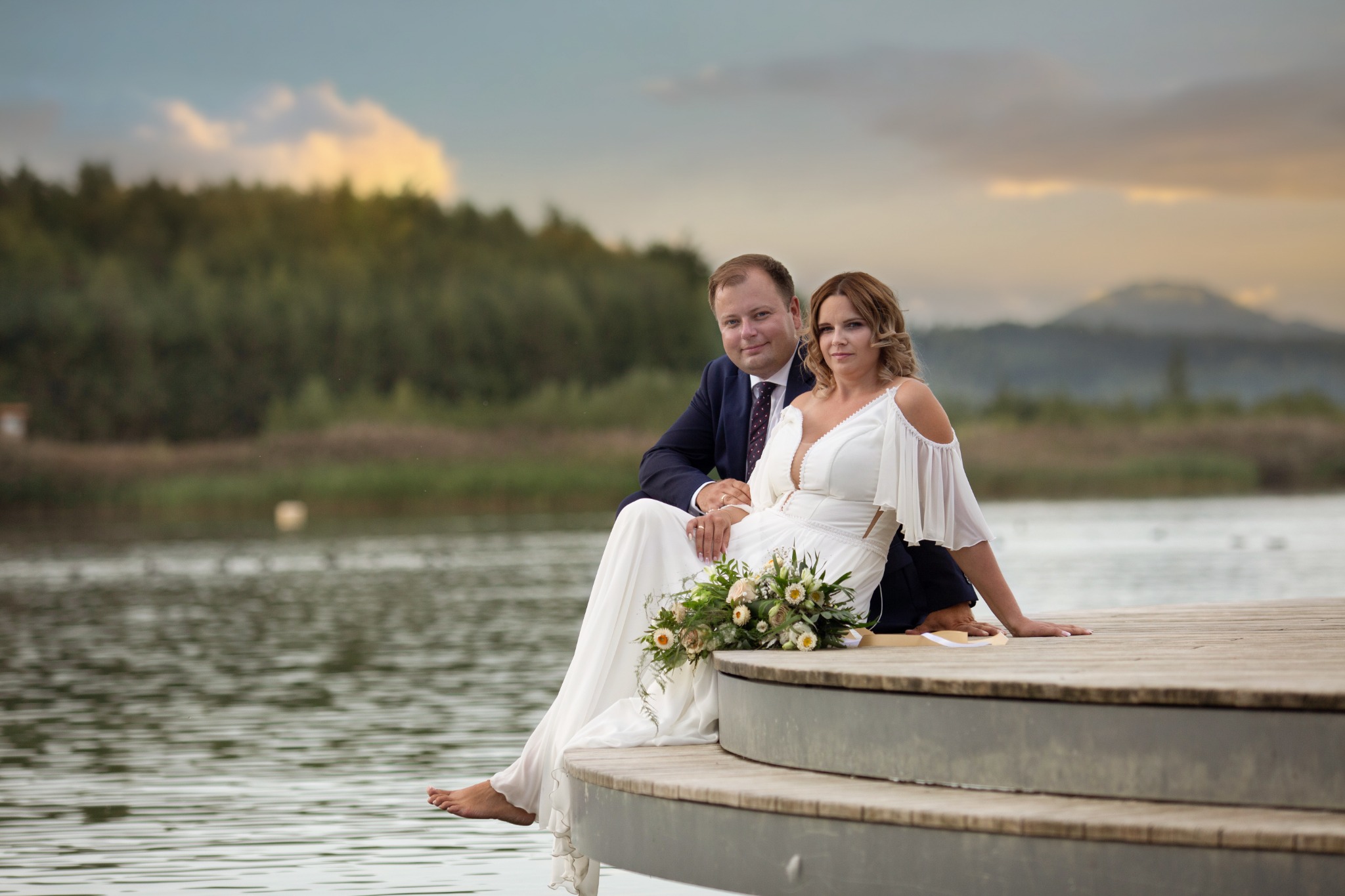 Para młoda siedząca na drewnianym pomoście nad jeziorem o zachodzie słońca, panna młoda w sukni z odkrytymi ramionami i bukietem polnych kwiatów, pan młody w granatowym garniturze.