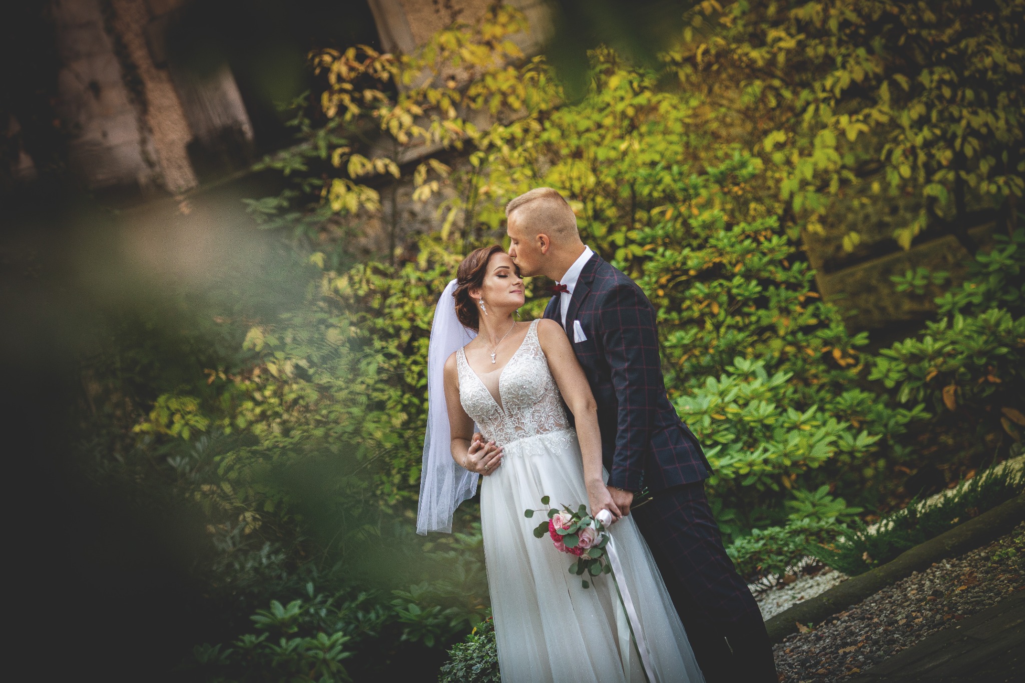 Panna młoda w koronkowej sukni i welonie trzyma bukiet różowych kwiatów, pan młody w bordowej marynarce całuje ją w czoło na tle zieleni.