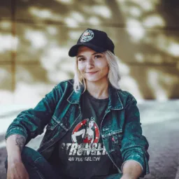 Uśmiechnięta blondynka w dżinsowej kurtce i czapce z daszkiem siedzi na tle złotej ściany. Na ręce widoczny tatuaż. Koszulka z motywem Wonder Woman.