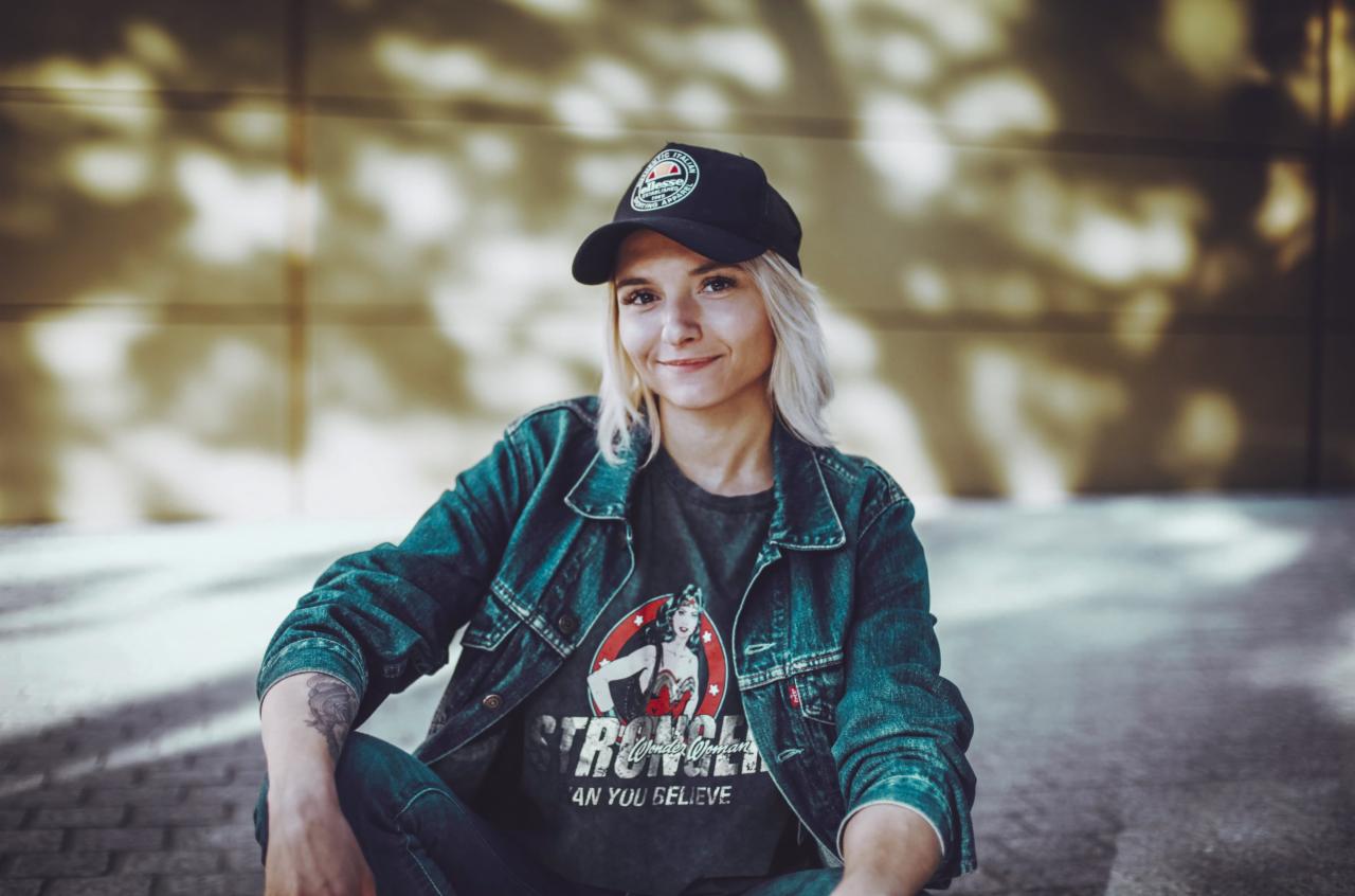 Uśmiechnięta blondynka w dżinsowej kurtce i czapce z daszkiem siedzi na tle złotej ściany. Na ręce widoczny tatuaż. Koszulka z motywem Wonder Woman.