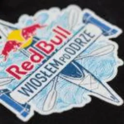 Zbliżenie na naszywkę z logo Red Bull Wiosłem po Odrze, przedstawiającą skrzyżowane wiosła i stylizowane fale, umieszczoną na czarnym materiale.