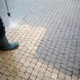 Tajfun - Czyszczenie kostki brukowej za pomocą myjki ciśnieniowej, widoczna różnica w kolorze między umytą a brudną powierzchnią, osoba w zielonych gumowcach obsługuje urządzenie.