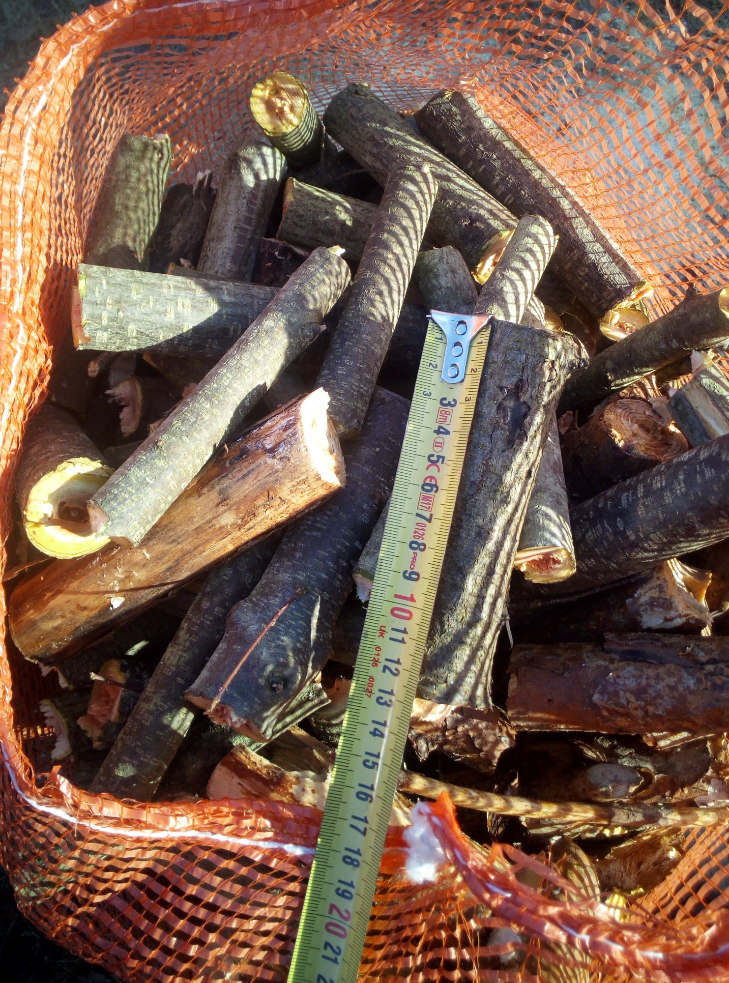 Kłody drewna kominkowego, pocięte na kawałki, w pomarańczowej siatce, obok miara zwijana pokazująca długość jednego z kawałków.