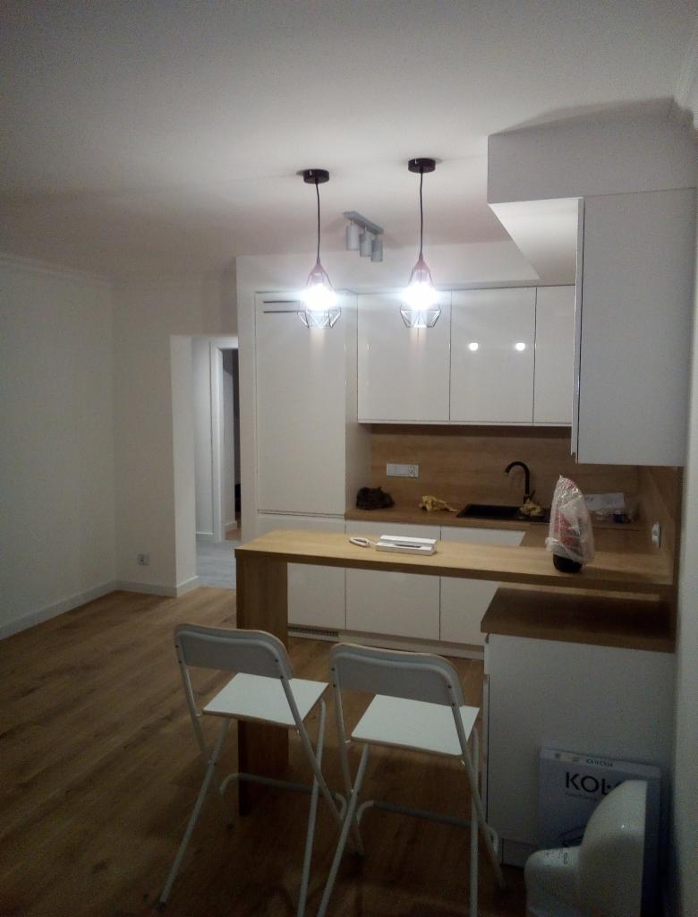 Nowoczesna kuchnia z białymi szafkami, drewnianym blatem i dwoma wiszącymi lampami w kształcie diamentów, widok z perspektywy dwóch białych krzeseł.