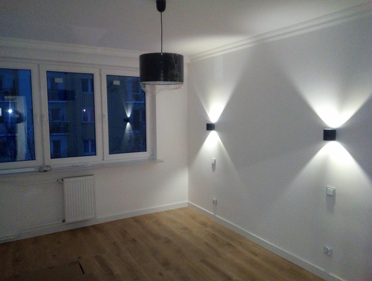 Wykończone wnętrze pokoju z parkietem, białymi ścianami, oknem z widokiem na budynek i nowoczesnym oświetleniem ściennym oraz wiszącą lampą z abażurem jeszcze w folii ochronnej.