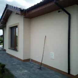 Wykończona elewacja domu jednorodzinnego w jasnym kolorze, widoczne okno z brązową ramą, czarna rynna, kostka brukowa na podjeździe i narzędzie oparty o ścianę.