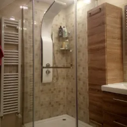 Nowoczesna kabina prysznicowa z hydromasażem, mozaikowe płytki w odcieniach beżu, drewniana szafka łazienkowa i grzejnik drabinkowy w jasnej łazience.