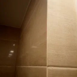 Beżowe, błyszczące płytki ceramiczne imitujące naturalny kamień, ułożone w rogu łazienki, widoczne fugi.