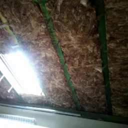 Izolacja poddasza wełną mineralną, widoczne krokwie z zielonymi łatami i okno dachowe z mocowaniem, oświetlone jasnym światłem.