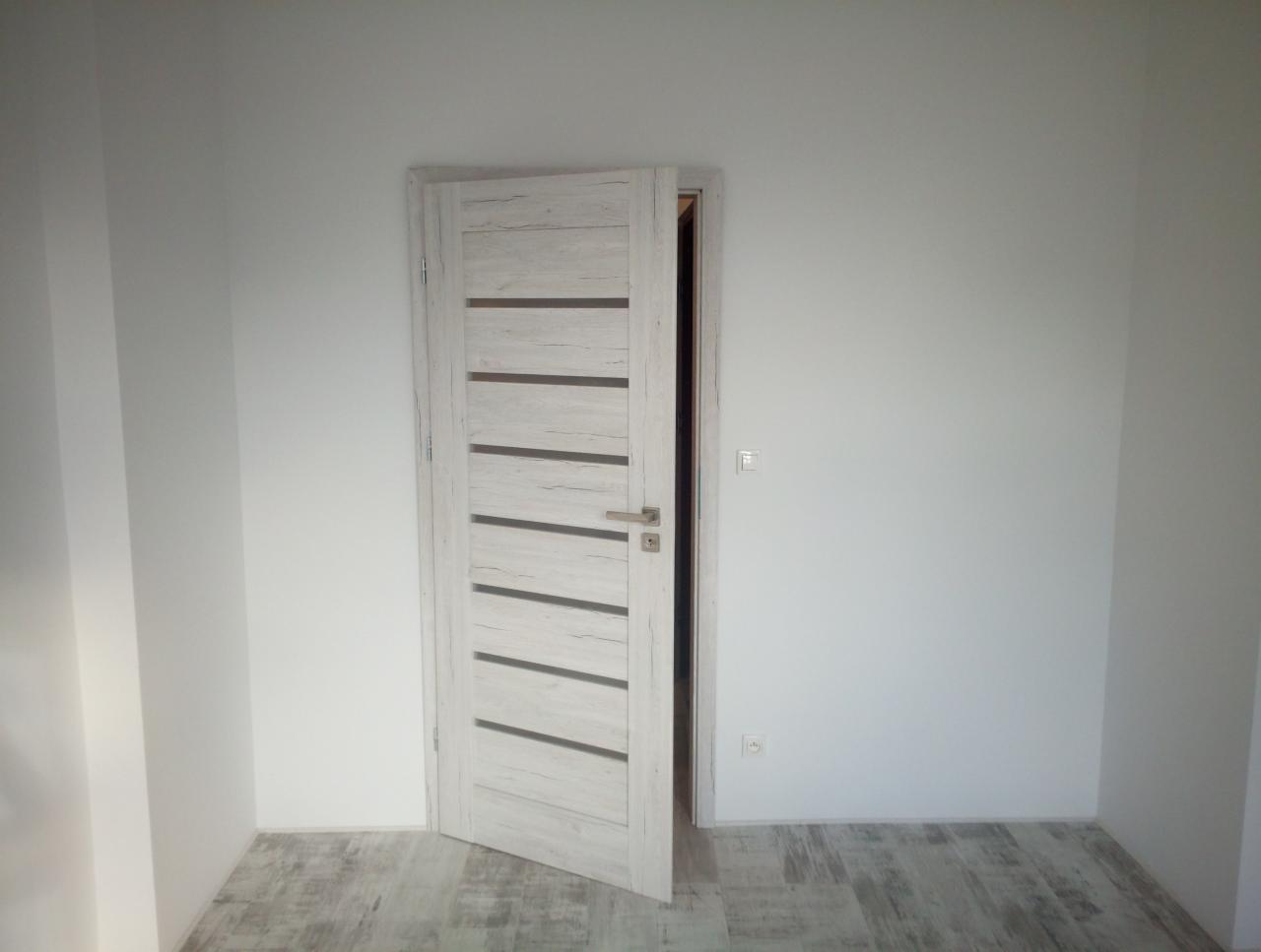 Otwarta, wewnętrzna, biała, drewnopodobna drzwi z poziomymi, ciemnymi paskami w nowo wyremontowanym, jasnym pokoju z panelami podłogowymi.